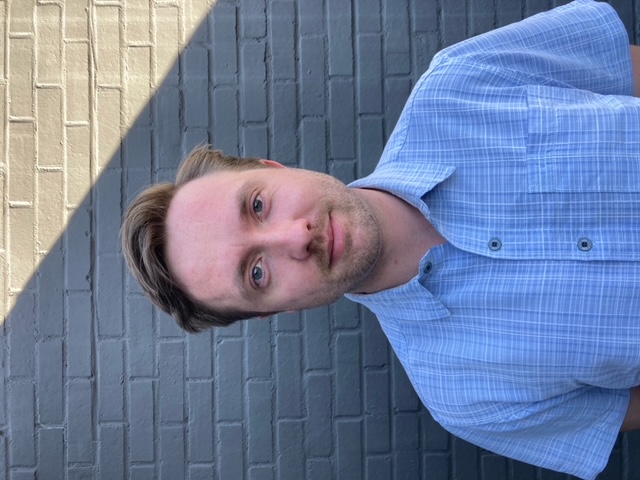 Neil Johnson stands in front of a gray brick wall, smiling and wearing a blue button down shirt.